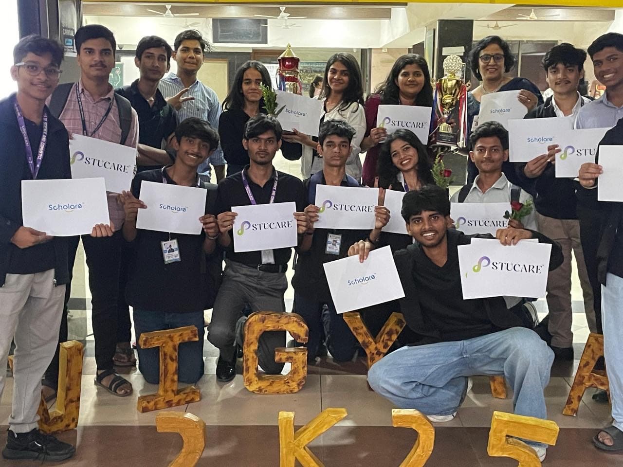 Students holding Scholare and Stucare cards at the JTGYA 2K25 college festival, with trophies displayed behind them